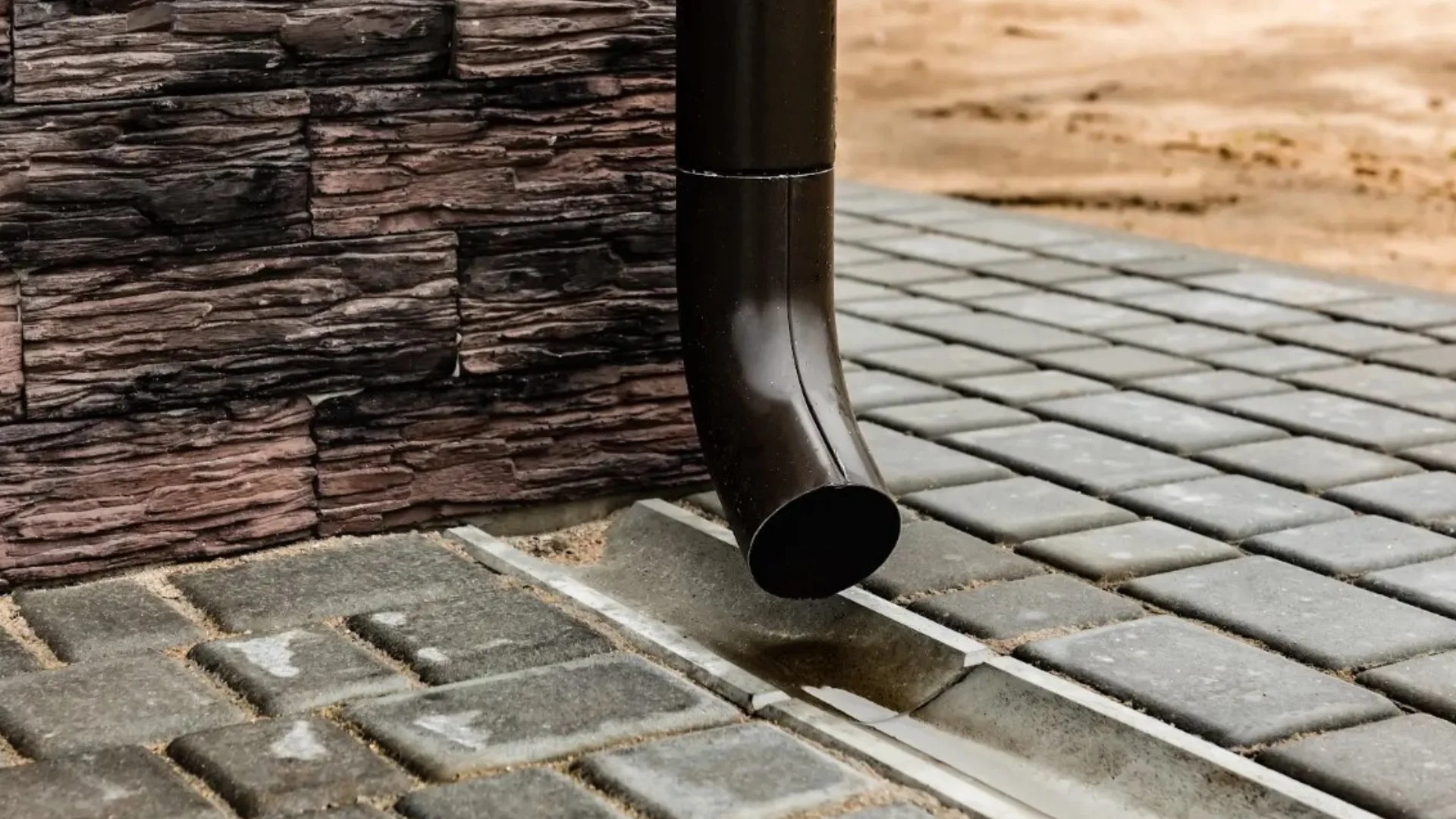 Close-up of a downspout draining water onto a paved area, with a decorative stone wall in the background.