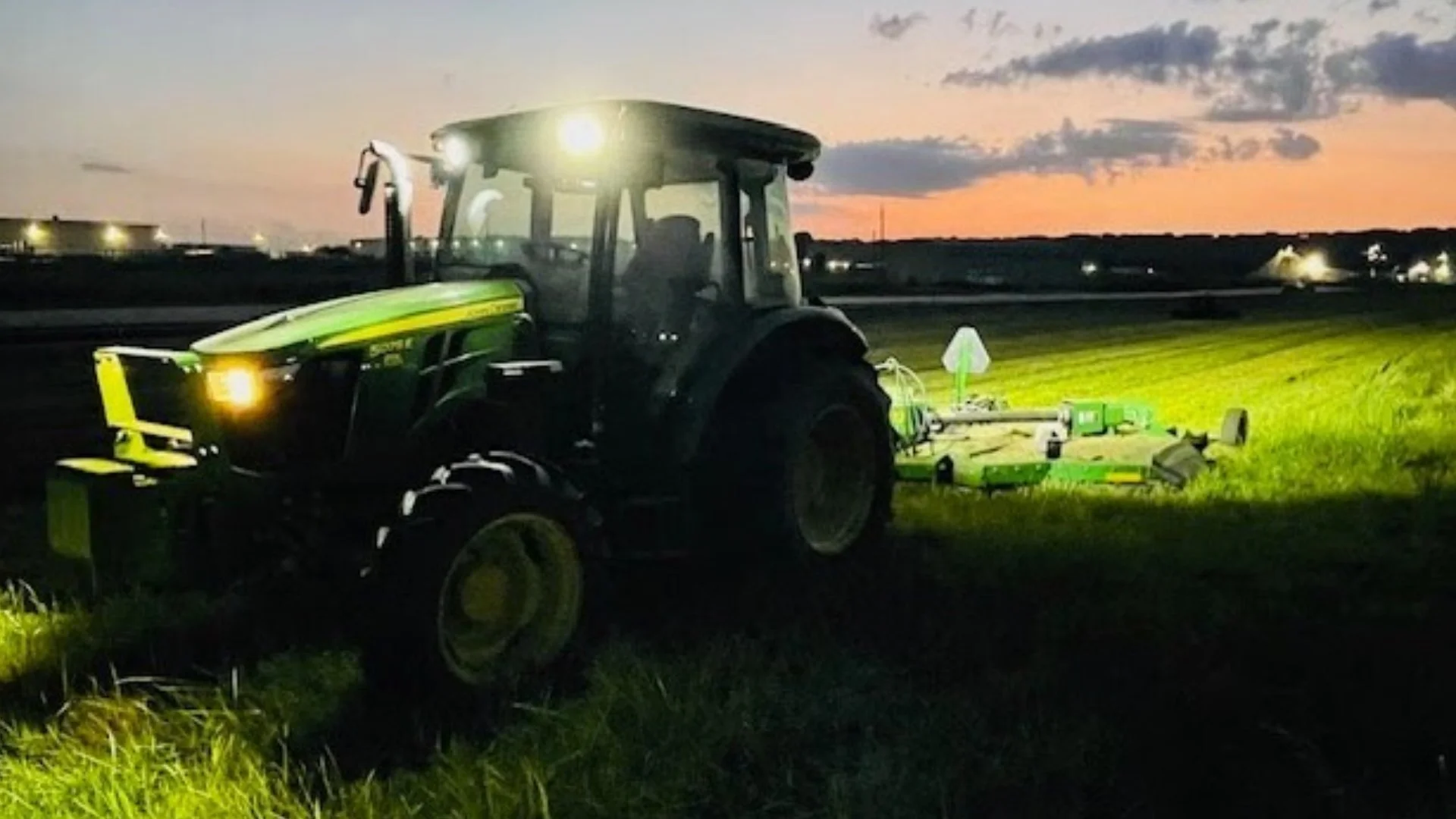 Tractor operating at dusk with headlights on, silhouetted against a sunset sky while mowing a field.